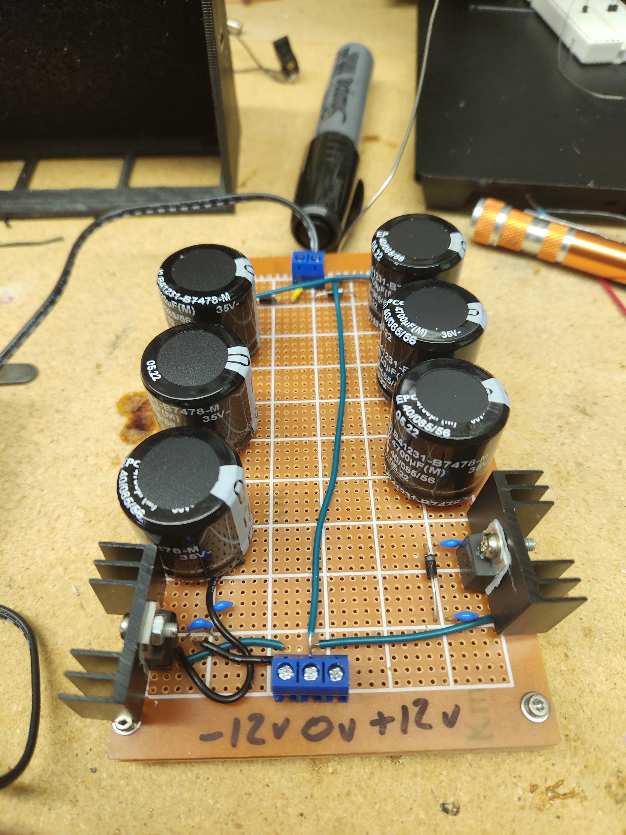 A photo of a homemade powersupply, sitting on a work bench.
                It is a large, rectangular, brown circuit board with six large
                capacitors and two voltage regulators with large aluminum heatsinks.
                There are three terminals on the front of the board labeled '-12V',
                '0V', and '+12V'. There are various tools scattered around.